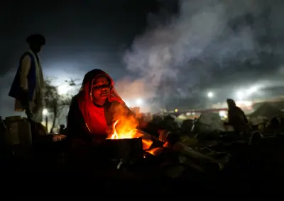 Rajasthani woman blowing air into a small bonfire at night to cook food during Pushkar Mela, her face lit by the warm glow of the fire.